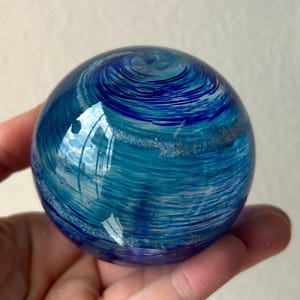 May include: A blue glass orb with white and blue swirls and flecks of silver. The orb is smooth and shiny.