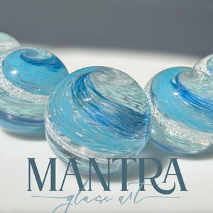 May include: Five spherical glass beads featuring swirling patterns of light blue, dark blue, and white with embedded silver glitter accents. The text MANTRA glass art is printed in dark blue at the bottom centre of the image below the glass beads.