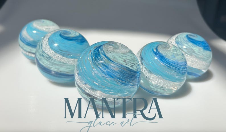 May include: Five spherical glass beads featuring swirling patterns of light blue, dark blue, and white with embedded silver glitter accents. The text MANTRA glass art is printed in dark blue at the bottom centre of the image below the glass beads.