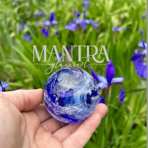 May include: A round, blue glass art piece with swirling white and silver accents. The glass sphere is held in a hand, with a blurred background of green foliage and purple flowers. The text "MANTRA glass art" is visible.