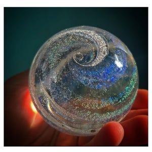 May include: A clear glass sphere with iridescent swirls of blue, green, and silver. The sphere is held in a hand, with a warm light emanating from below. The design resembles a galaxy or nebula.