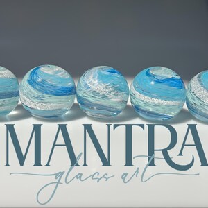 May include: Five spherical glass paperweights arranged in a row, each featuring swirling patterns of light blue, dark blue, and white with embedded silver glitter. The text MANTRA glass art is printed in dark blue below the glass spheres.