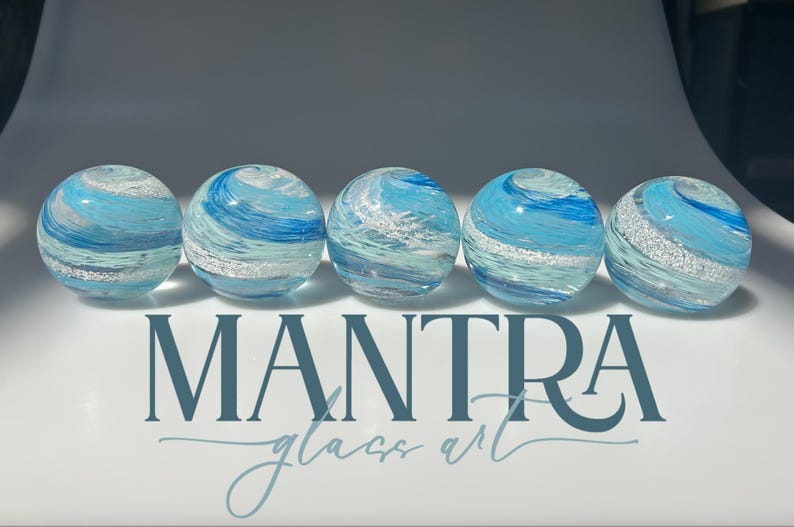 May include: Five spherical glass paperweights arranged in a row, each featuring swirling patterns of light blue, dark blue, and white with embedded silver glitter. The text MANTRA glass art is printed in dark blue below the glass spheres.