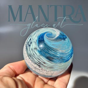 May include: A spherical glass paperweight featuring a swirling pattern of deep blue, light blue, and white colours. The surface displays a textured, silver-toned band encircling the lower portion, with the text MANTRA glass art visible above.