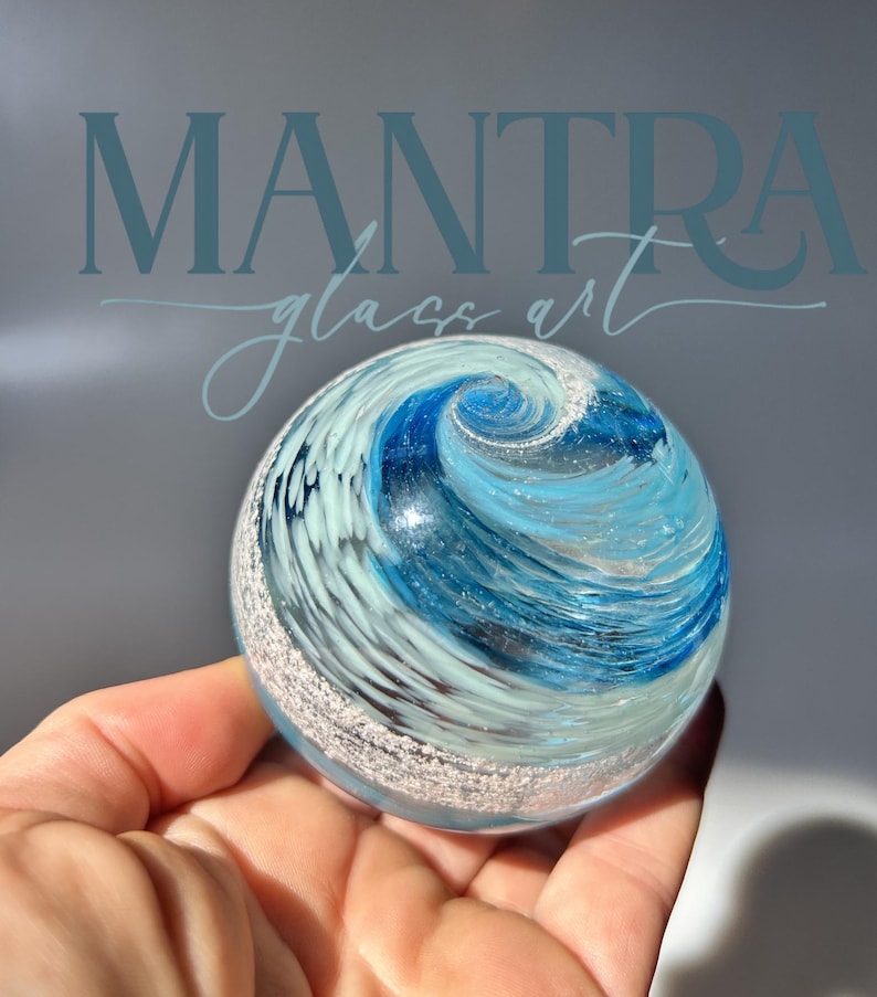 May include: A spherical glass paperweight featuring a swirling pattern of deep blue, light blue, and white colours. The surface displays a textured, silver-toned band encircling the lower portion, with the text MANTRA glass art visible above.