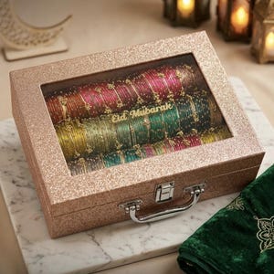May include: A rectangular rose gold glitter storage case with a clear glass lid featuring the text Eid Mubarak. Inside, rows of multi-colored bangles in shades of red, green, yellow, and blue are arranged, secured by a silver metal latch and handle.