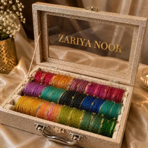 Pode incluir: Uma caixa de armazenamento retangular dourada e brilhante com uma tampa de vidro transparente com o texto ZARIYA NOOR. A caixa contém várias fileiras de pulseiras metálicas finas em várias cores, incluindo vermelho, laranja, roxo, azul-petróleo, preto, azul e verde.