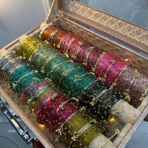 May include: A collection of multi-coloured glass bangles arranged in rows within a textured, metallic-toned storage case. The bangles feature translucent hues of yellow, green, pink, and dark grey, each adorned with small, metallic decorative accents.