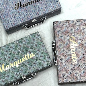 Op de afbeelding: Drie rechthoekige koffers met glinsterende, veelkleurige texturen en zilveren metalen handgrepen. Elke koffer is voorzien van een gepersonaliseerde naam in goudkleurige schrijfletters: Hannah, Marquetta en Annie op de deksels.