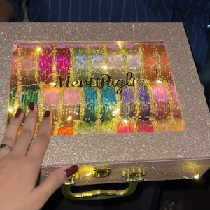 Könnte beinhalten: Eine rechteckige, glitzernde champagnerfarbene Aufbewahrungsbox mit transparentem Deckel, die Reihen von bunten Armreifen enthält. Der Schriftzug "Meri Pagli" ist in schwarzer Schrift auf dem Fenster gedruckt. Goldfarbener Metallgriff.