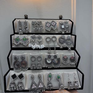 Könnte beinhalten: Ein schwarzer Schmuckständer mit sechzehn Paar silberfarbenen Ohrringen. Die Kollektion umfasst verschiedene Designs, darunter Jhumkas, geometrische Formen, hängende Anhänger sowie Akzente in grüner, rosa und weißer Emaille.