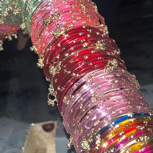Puede incluir: Una colección de brazaletes circulares y translúcidos apilados en un degradado de colores que incluye verde claro, rosa brillante, rojo intenso, rosa suave, azul y amarillo. Cada brazalete presenta pequeños adornos metálicos dorados.