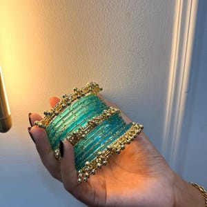 Könnte beinhalten: Ein Set aus gestapelten Armreifen mit durchscheinenden türkisfarbenen Glasröhren, die mit metallisch goldfarbenen Perlen verziert sind. Die kreisförmigen Armbänder sind in einem Bündel angeordnet und kombinieren glattes, leuchtend blaugrünes Glas mit strukturierten Goldmetalldetails.