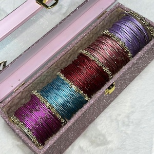 May include: A rectangular pink glittery storage box with a clear lid containing five stacks of thin metal bangles. The bangles are arranged in colour order: purple, light pink, deep red, bright blue, and magenta, each accented with gold beads.