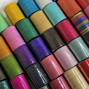 Può includere: Una collezione di numerosi braccialetti sottili in metallo disposti in file, caratterizzati da un'ampia varietà di colori solidi tra cui giallo, blu, verde, rosa, viola, arancione, argento, oro e nero, con una finitura testurizzata su ogni bracciale.