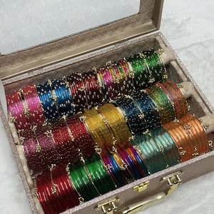 Op de afbeelding: Een collectie veelkleurige glazen armbanden gerangschikt in een rechthoekige, glitterende beige opbergdoos met een doorzichtig deksel. De armbanden hebben diverse tinten, waaronder rood, groen, blauw, geel, oranje en roze, elk versierd met kleine metalen accenten.