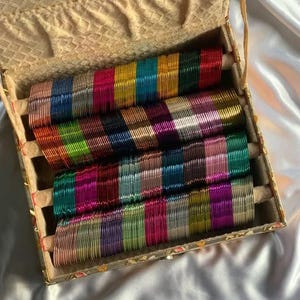 Könnte beinhalten: Eine Sammlung dünner, metallischer Armreifen in einem breiten Spektrum leuchtender Farben, darunter Rot, Blau, Grün, Gold, Silber und Pink, angeordnet in Reihen auf vier horizontalen, gepolsterten Stangen in einer rechteckigen Schmuckschatulle.