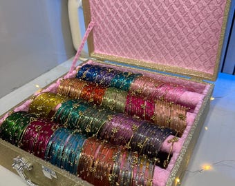 May include: A gold glittery storage case with a pink textured interior lining, containing multiple rows of thin, translucent bangles in various colors including green, red, blue, purple, and gold, each featuring small gold-toned metallic accents.