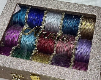 May include: A rose gold glitter storage case with a clear lid and gold handle, containing rows of multi-colored metallic bangles. The name Annie is written in gold script on the glass lid. The bangles feature various colors including blue, silver, purple, red, and gold.