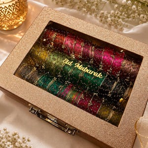 Könnte beinhalten: Eine rechteckige, glitzernde goldene Aufbewahrungsbox mit einem klaren Glasdeckel, die Reihen dünner, metallischer Armreifen in Rot-, Grün- und Schwarztönen enthält. Auf dem Glasdeckel befindet sich der goldene Schriftzug Eid Mubarak.