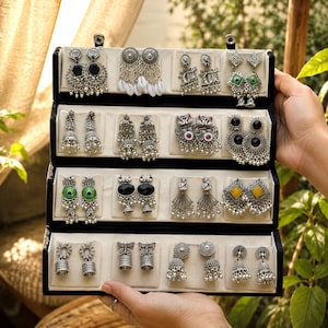 Può includere: Un espositore per gioielli nero contenente sedici paia di orecchini color argento. La collezione presenta vari design, tra cui jhumka, orecchini pendenti con pietre colorate, dettagli in conchiglia e intricati motivi in metallo.
