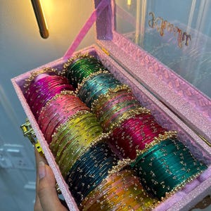 Könnte beinhalten: Eine rechteckige, glitzernde rosa Aufbewahrungsbox mit transparentem Deckel, die mehrere Reihen bunter Armreifen enthält. Die Armreifen sind durchscheinend und mit kleinen goldfarbenen Metallakzenten versehen. Die Box hat einen goldenen Verschluss.