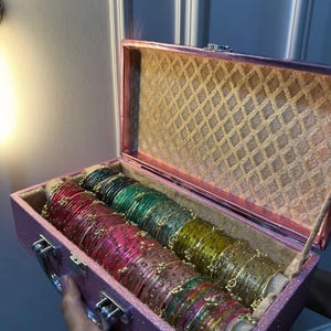 May include: A rectangular pink glittery storage box with a hinged lid, containing multiple rows of thin, metallic bangles in various colors including red, green, yellow, and pink, each adorned with small, delicate gold-toned decorative accents.