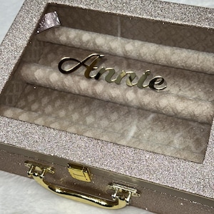 Op de afbeelding: Een rechthoekige sieradendoos met een roségouden glitterafwerking en een doorzichtig glazen deksel. De naam Annie staat in gouden schrijfletters op het glas. De doos is voorzien van een gouden metalen handvat en een gouden sluiting.