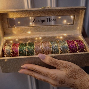 May include: A rectangular, glittery gold storage box with a clear window displaying the text Zariya Noor, filled with a row of multicolored bangles in shades of silver, pink, green, gold, blue, and purple, each featuring small gold bead accents.