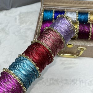 Op de afbeelding: Een stapel dunne metalen armbanden in paarse, rode en blauwe tinten, elk met een textuur met glitterafwerking en geaccentueerd met kleine goudkleurige kralenringen. De armbanden zijn in een lange kolom gerangschikt, met een opbergdoos op de achtergrond.