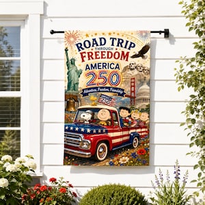 May include: A vertical garden flag featuring a red, white, and blue pickup truck with Peanuts characters, the Statue of Liberty, Mount Rushmore, and the text ROAD TRIP THROUGH FREEDOM AMERICA 250 Adventure, Freedom, Friendship, hanging on a black rod.