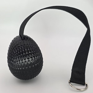 May include: Black textured egg-shaped massage ball with a black strap and a silver metal ring. The ball has a diamond pattern and is designed for muscle relief and self-massage.