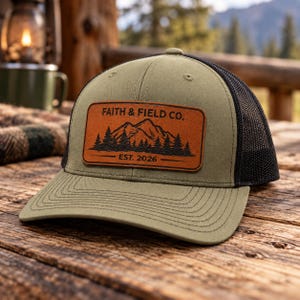May include: An olive green baseball cap with a black mesh back and a rectangular brown leather patch on the front. The patch features a black graphic of mountains and pine trees with the text FAITH & FIELD CO. and EST. 2026 printed on it.