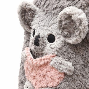 K&ouml;nnte beinhalten: Ein kuscheliges graues Koala-B&auml;r-Spielzeug mit einer rosa Tasche. Der Koala hat gro&szlig;e schwarze Augen, eine schwarze Nase und kleine schwarze Pfoten. Die weiche, pelzige Textur des Koalas ist sichtbar.