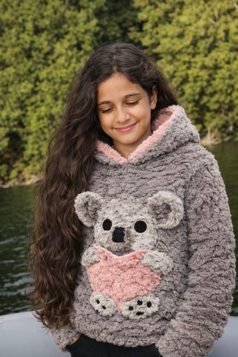 K&ouml;nnte beinhalten: Grauer Kapuzenpullover mit Koala-B&auml;r-Design. Der Koala hat gro&szlig;e schwarze Augen, eine schwarze Nase und eine herzf&ouml;rmige rosa Tasche. Die Kapuze ist mit rosa Stoff gef&uuml;ttert. Der Pullover ist aus weichem, pl&uuml;schigem Material.