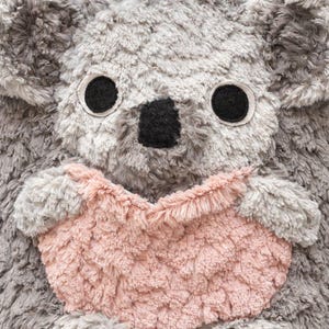 K&ouml;nnte beinhalten: Ein Pl&uuml;sch-Koala mit grauem Fell, gro&szlig;en schwarzen Augen und einer schwarzen Nase. Der Koala h&auml;lt ein herzf&ouml;rmiges Objekt in Rosa. Das Spielzeug hat ein weiches, strukturiertes Aussehen, was auf ein kuscheliges Design hindeutet.
