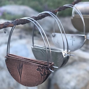 Peut inclure: Trois paires de boucles d'oreilles créoles en argent avec un design en demi-cercle. Les boucles d'oreilles sont suspendues à un fil rouillé. La boucle d'oreille du bas est en cuivre avec un motif floral.