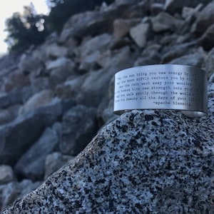 May include: A silver metal bracelet with an engraved quote: "May the sun bring you new energy by day, the moon softly restore you by night, may the rain wash away your worries, may the breeze blow new strength into your soul, may you walk gently through the world and see its beauty all the days of your life." - Apache Blessing