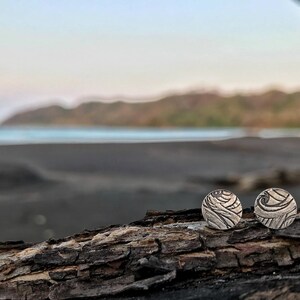 May include: Two silver earrings with a textured design resembling waves. The earrings are on a piece of driftwood with a blurred background of a beach and ocean.