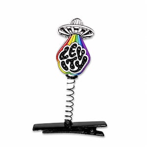 May include: A black metal hair clip featuring a spring-mounted charm of a silver UFO emitting a rainbow-colored tractor beam. The beam contains the word LEVITY written in white, stylized, bubbly letters against a black, star-speckled background.