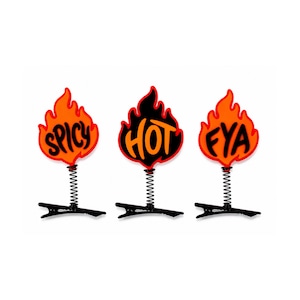 May include: Three novelty clips with flame-shaped designs. Each flame is orange and red, with the words "SPICY", "HOT", and "FYA" in black. The flames are attached to a black clip via a silver spring.