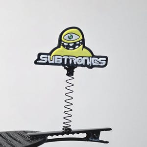 May include: A hair clip featuring a yellow, one-eyed monster character with a black and white toothy grin. The character is attached to a black metal spring mounted on a black alligator hair clip. The text SUBTRONICS is printed in white.