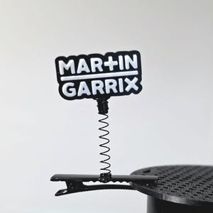 May include: A black and white clip featuring the text "MAR+IN GARRX". The clip has a spring and is attached to a black base. The text is in a bold, sans-serif font.