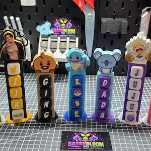 Puede incluir: Siete soportes de exhibición verticales impresos en 3D con varios personajes en la parte superior, incluyendo Zoro, una figura sin camisa, Shooky, Squirtle, Koya, Luffy y Hello Kitty. Cada soporte muestra bloques de letras apilados que deletrean los nombres CHAOS, MIJK, GING, KOBE, DADA, JUJU y LETICIA.