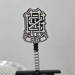 Puede incluir: Un pin de metal negro y plateado con un logotipo estilizado construido a partir de formas que recuerdan a huesos. El diseño incluye una pancarta en la parte inferior con el texto GRIZ en letras negras. El pin está unido a un resorte metálico fino y un clip negro.