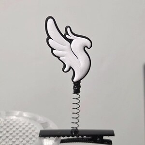 Può includere: Una molletta per capelli con una base in metallo nero a coccodrillo e una sottile molla metallica verticale fissata a un emblema bianco a forma di ala con un bordo nero. L'emblema raffigura un'ala di uccello stilizzata rivolta verso l'alto.