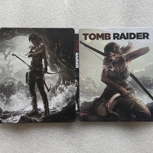 May include: Two metal game cases featuring the title "TOMB RAIDER." One case shows a woman with a bow and arrow standing in front of a stormy background. The other case shows a woman kneeling with a bow and arrow.