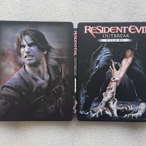May include: Resident Evil Outbreak File #2 game case. The case is open, revealing the front and spine. The front cover features a man with an angry expression. The back cover shows a person's hands grabbing a zombie's leg.