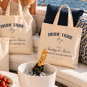 May include: Cream-colored canvas tote bags with the text "BRIDE TRIBE" and "Toasting to Forever" in navy blue. A champagne bottle in an ice bucket is in the foreground. The bags are for a bachelorette party in Cabo San Lucas.