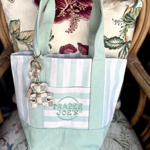 May include: A light green and white striped canvas tote bag featuring the embroidered text Trader Joe's in a circular design on the front pocket. A small, checkered bear-shaped keychain with a tassel is attached to the handle of the bag.